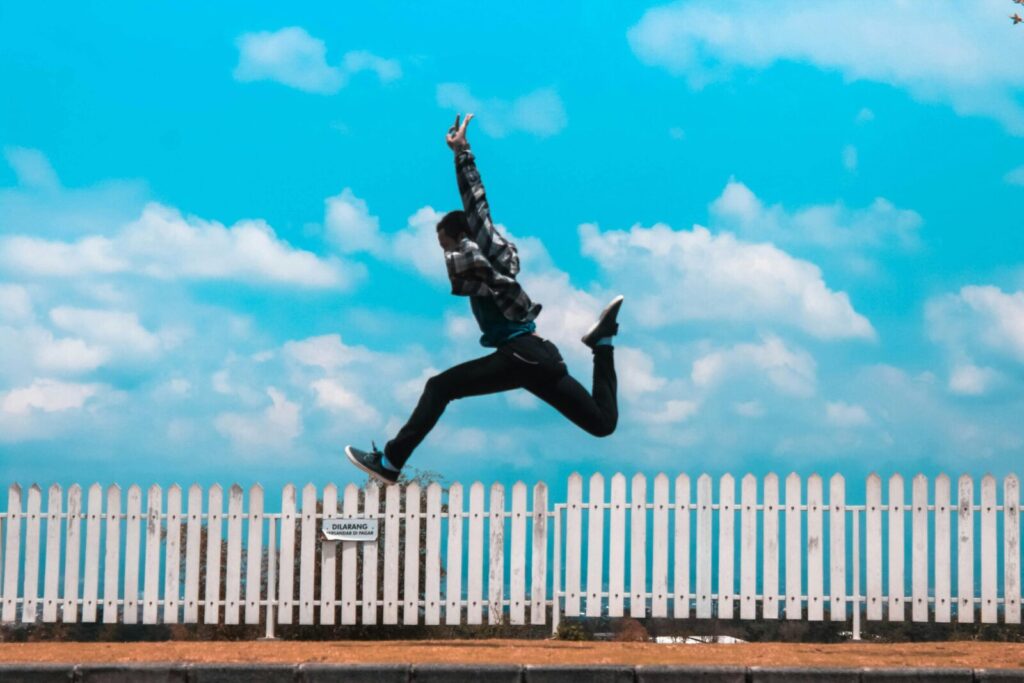 Mid-air capture of a person leaping in front of a picket fence under a bright blue sky.